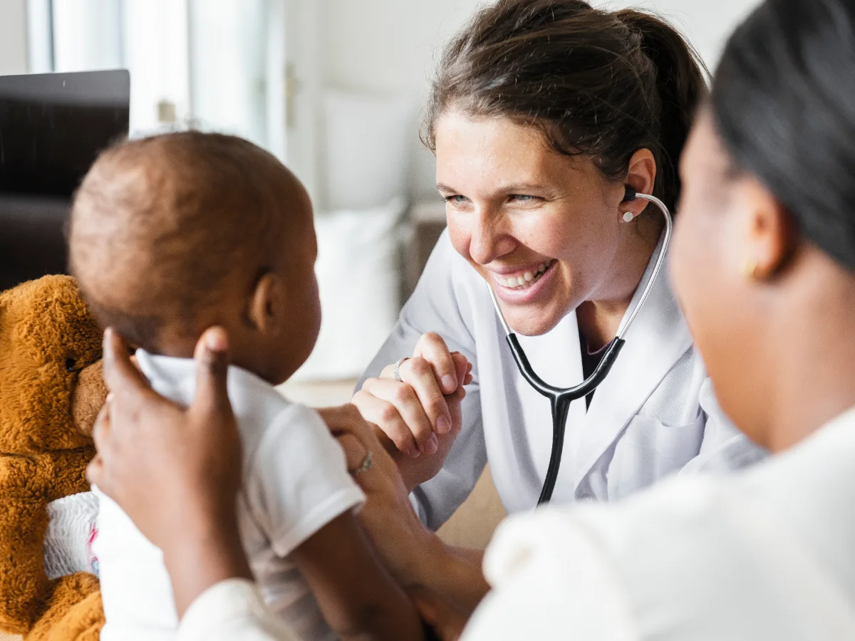 Médica examina bebê que segura um ursinho, com a mãe ao lado — imagem ilustrativa